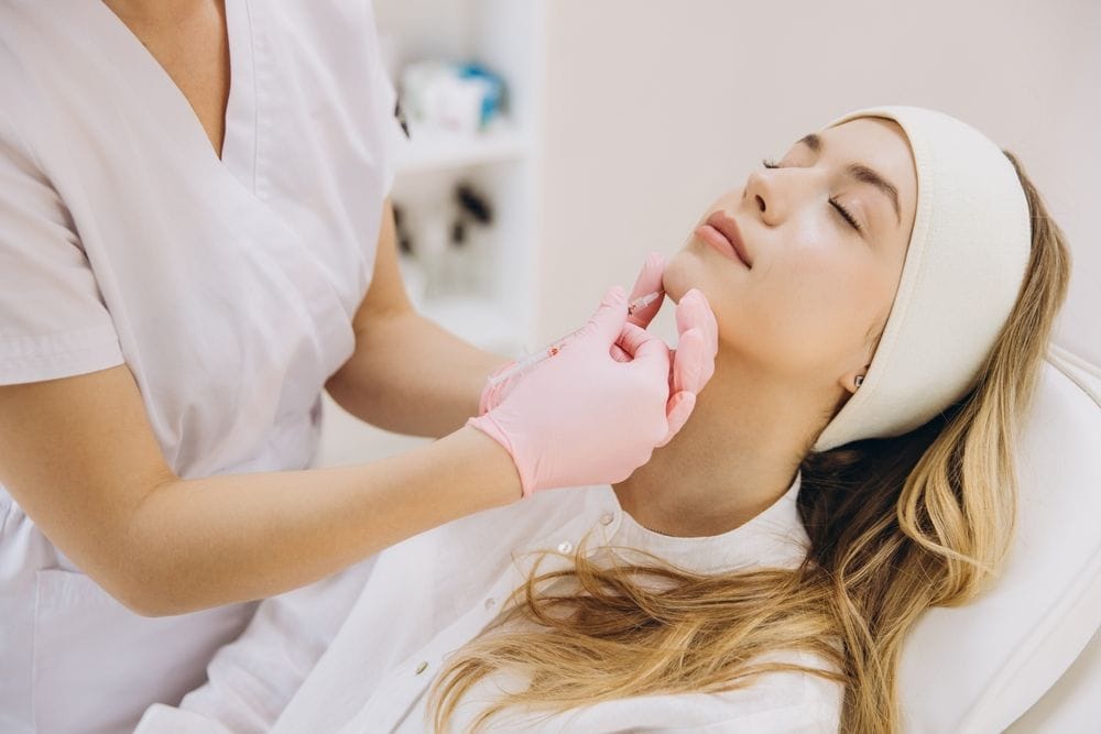 A beautician administering a dermal filler to a patient’s chin.
